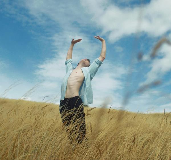 A man in an open blue shirt stretches his arms above him in a field of golden corn against a blue sky
