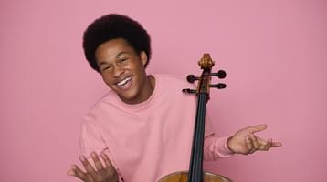 The cellist pictured smiling against a pink backdrop, wearing a t-shirt in the same shade of pink sitting behind his cello.