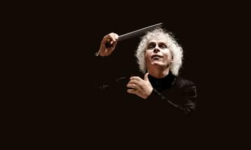 A man with curly white hair wearing all black against a black background, holding a conductor's baton aloft
