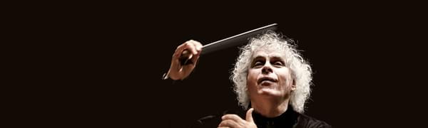 A man with curly white hair wearing all black against a black background, holding a conductor's baton aloft