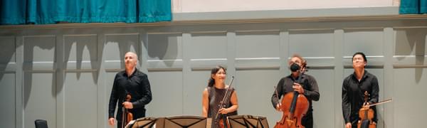 Four string players stand in front of an applauding audience against a light blue wall