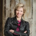 A blonde woman in a black leather jacket stands with her arms crossed. She is smiling.