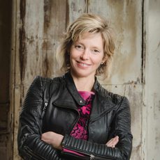 A blonde woman in a black leather jacket stands with her arms crossed. She is smiling.