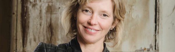 A blonde woman in a black leather jacket stands with her arms crossed. She is smiling.