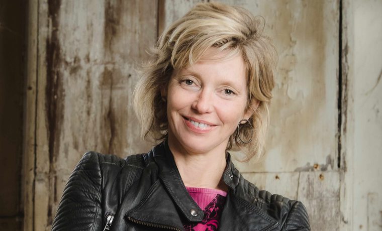 A blonde woman in a black leather jacket stands with her arms crossed. She is smiling.