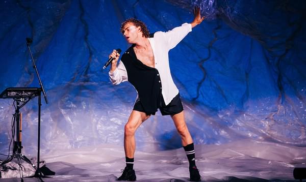 A young man dressed in a half-white, half-black shirt, black shorts and boots leans forward and sings into a microphone onstage, with one arm stretched out behind him.