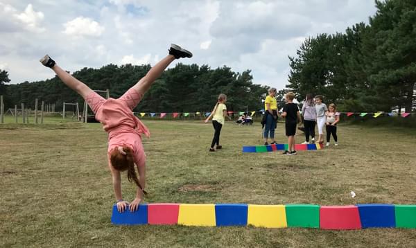 A girl in a pink playsuit cartwheels over a row of colourful blocks, with children playing in the background.