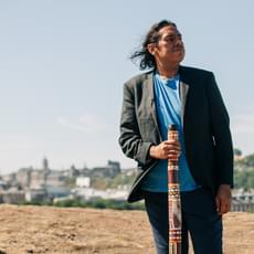 An aboriginal man in a bright blue tshirt and black blazer stands holding a didgeridoo with a view of Edinburgh behind him