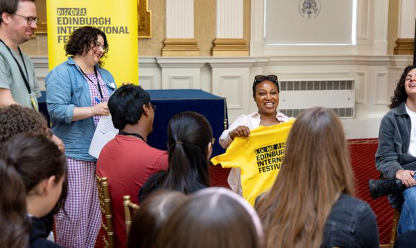 A Black woman smiles and holds up a yellow Edinburgh International Festival t-shirt, while an audience watches and smiles.