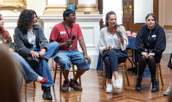 A White woman speaks into a microphone. Sat next to her are teenagers who smile and listen.