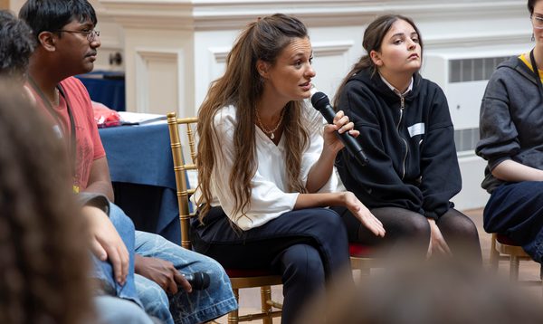 A White woman in a white blouse speaks into a microphone. Teenagers sit around her, listening.