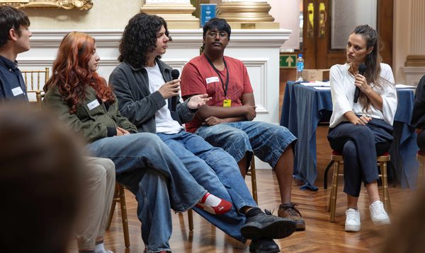 A teenager with long dark hair and a microphone speaks to a woman sat across from him. Teenagers watch and listen.