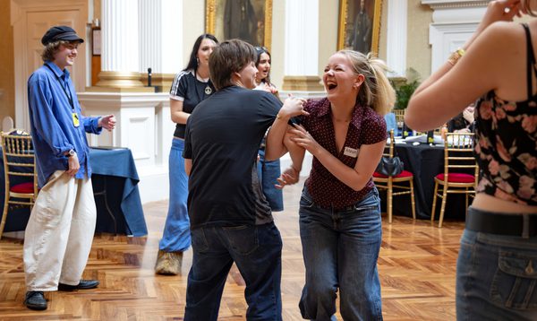 Two teenagers dance arm in arm, one of them is laughing. Others stand and watch, smiling.