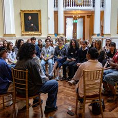A group of teenagers sit gathered to listen to speakers with their back to the camera.