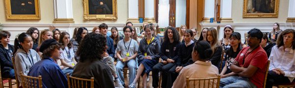 A group of teenagers sit gathered to listen to speakers with their back to the camera.