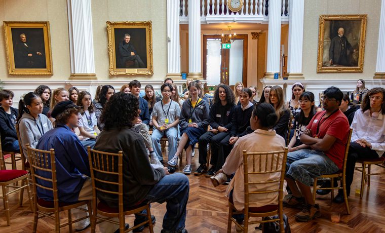 A group of teenagers sit gathered to listen to speakers with their back to the camera.