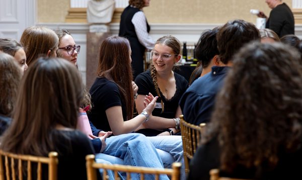 Amidst a crowd of people sitting, two teenagers chat excitedly.
