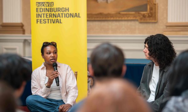 A Black woman speaks into a microphone with a Edinburgh International Festival banner behind her. People sit around her, listening,