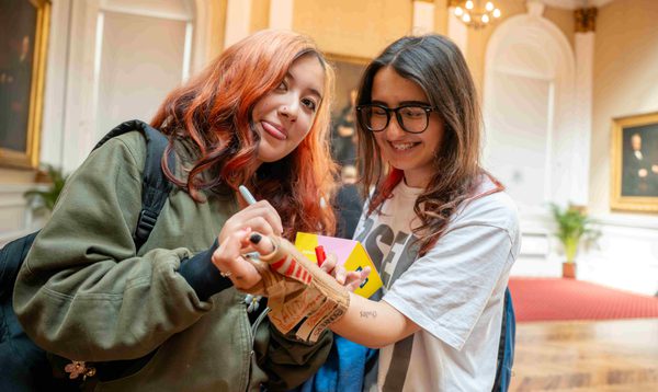 A teenage girl with dyed orange hair signs the arm brace of her friend while she sticks her tongue out at the camera.