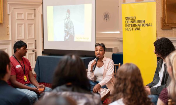 A Black woman speaks to a teenage audience, with a projection of her posing with a saxophone in the background.