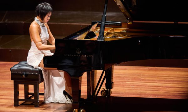 A woman with short, dark hair, wears a white dress while playing piano