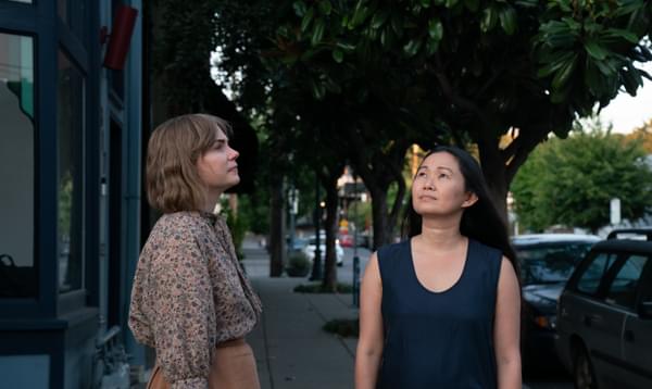Two women stand in the street. One smiles and looks at the sky. The other gazes off into the distance.