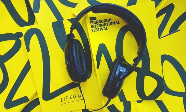 A pair of black over-ear headphones photographed lying on top of a scattered stack of yellow 2018 Edinburgh International Festival brochures.